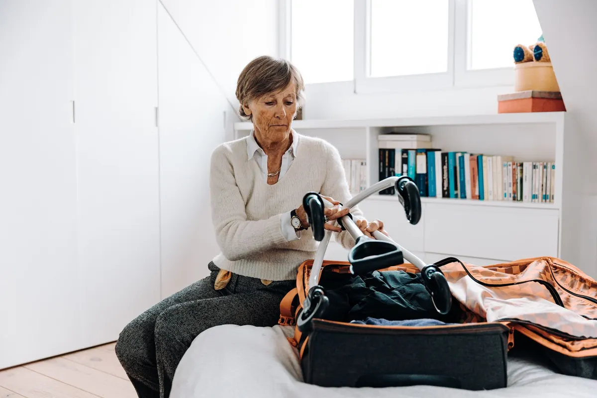 Eine ältere Frau packt Kleidung neben ihrer Gehhilfe in einen Koffer